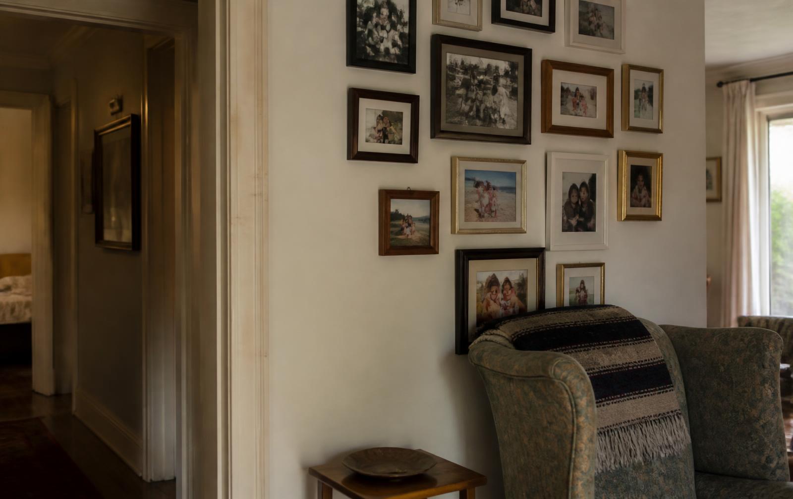 A lived-in family home interior: an entry hallway opening onto a living room, a wall of framed family photographs in mismatched frames, a worn armchair with a folded blanket and soft afternoon light