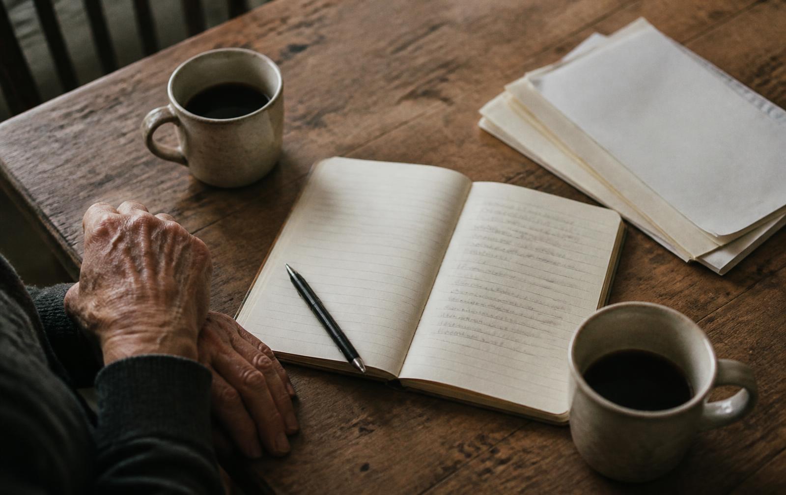 An overhead view of a wooden kitchen table in morning light with two coffee cups, an open notebook, a pen and a small stack of papers