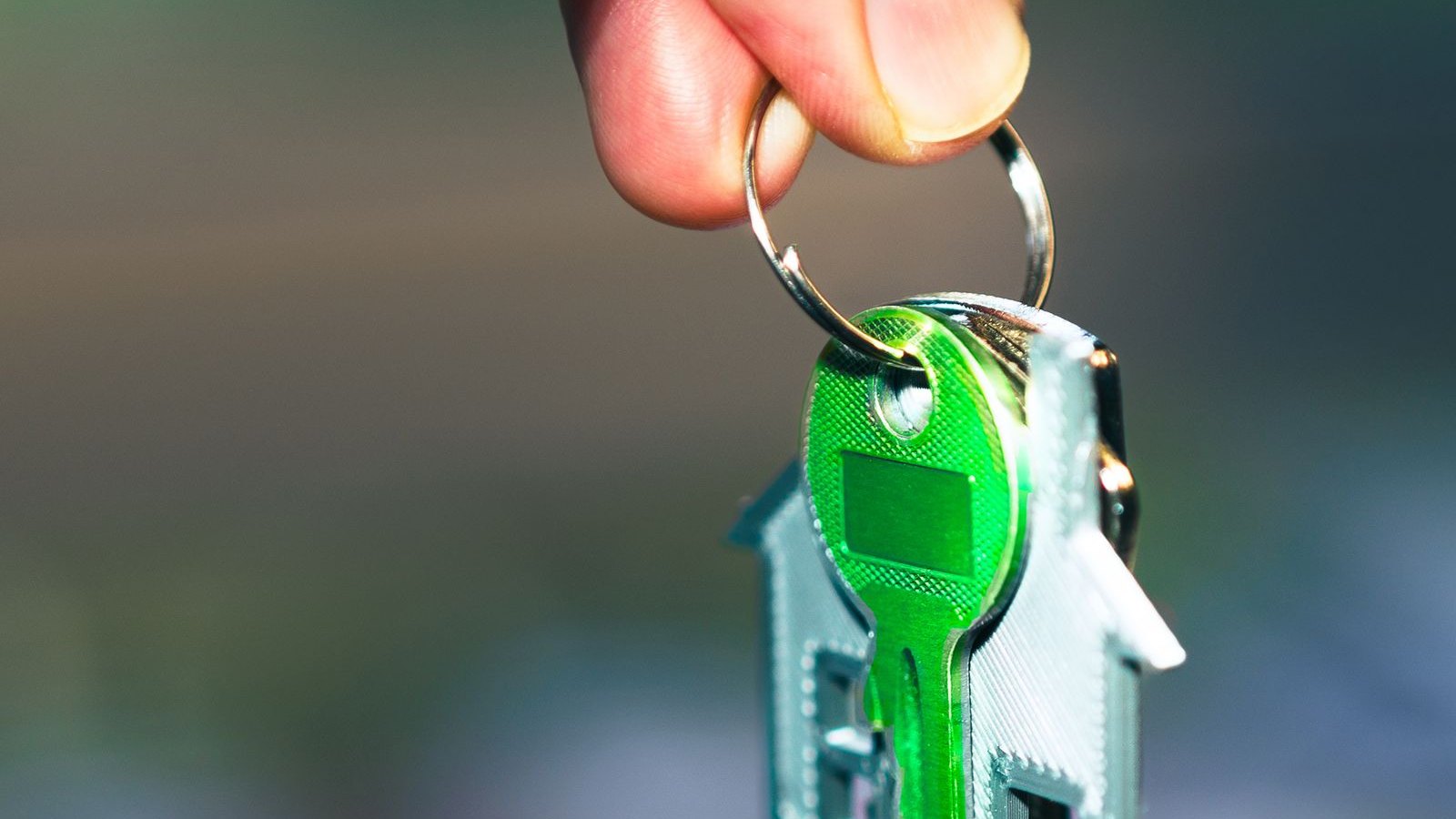 A hand holding a house key on a green house-shaped keyring in sunlight.